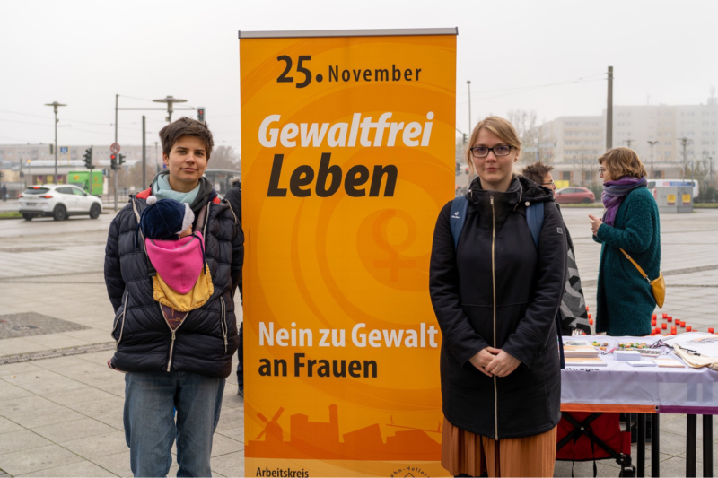 Zwei Frauen stehen neben einem orangefarbenen Banner mit der Aufschrift: „25. November: Gewaltfrei leben. Nein zu Gewalt an Frauen.“ Im Hintergrund ist der Alice-Salomon-Platz in Berlin-Hellersdorf zu sehen.
