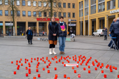 Zwei Frauen stehen auf dem Alice-Salomon-Platz in Berlin-Hellersdorf vor einer Vielzahl an Kerzen in roten Glasbehältnissen. Die Kerzen dienen als Zeichen des Gedenkens