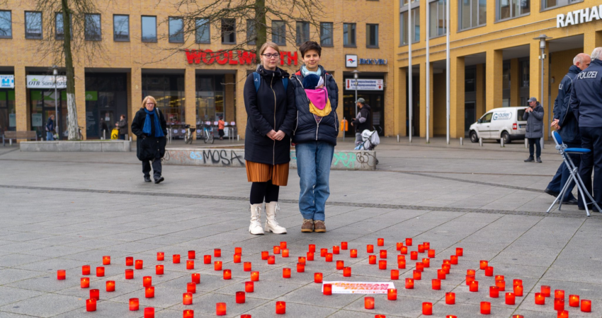 Zwei Frauen stehen auf dem Alice-Salomon-Platz in Berlin-Hellersdorf vor einer Vielzahl an Kerzen in roten Glasbehältnissen. Die Kerzen dienen als Zeichen des Gedenkens