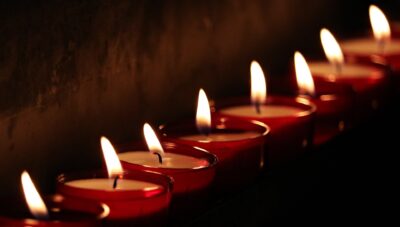 Mehrere kleine Kerzen, die in einer Reihen stehen, leuchten. Die Kerzen stehen in kleinen roten Gläsern. Der Rest des Bildes ist dunkel.