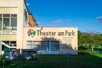 Theater am Park, im Hintergrund der blaue Himmel unigrüne Bäume.