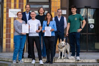 Vertreter*innen von Bündnis 90/Die Grünen und der CDU aus Marzahn-Hellersdorf stehen gemeinsam auf der Treppe vor dem Rathaus Marzahn. In der Hand halten sie eine unterschriebene Vereinbarung. Zu sehen sind: Chantal Münster, Christian Gräff, Max Linke, Maren Tepper, Nadja Zivkovic, Johannes Martin, Pascal Grothe und Hündin Susi.