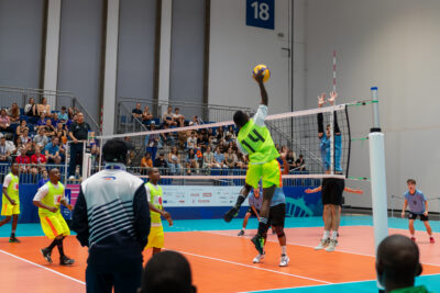 Zwei Mannschaften spielen Volleyball in einer Halle. Im Hintergrund sitzt Publikum