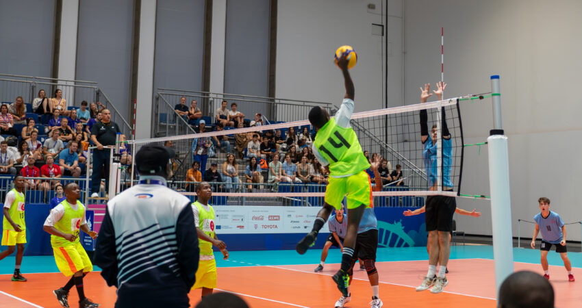 Zwei Mannschaften spielen Volleyball in einer Halle. Im Hintergrund sitzt Publikum