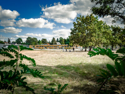 eine große Grünfläche und im Hintergrund ein Spielplatz mit spielenden Kindern.