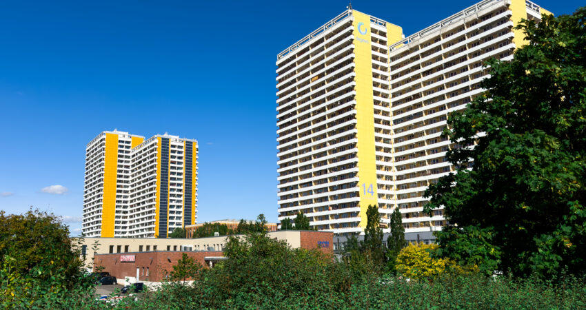 Zwei Hochhäuser. Im Hintergrund blauer Himmel und im Vordergrund grüne Bäume und Hecken.