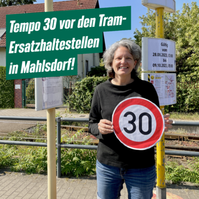 Die Grünen-Sprecherin Maren Tepper steht an einer Ersatzhaltestelle am Hultschiner Damm. Sie lächelt und hält ein Schild für Tempo 30 in der Hand. Neben ihr ist folgender Text eingeblendet: „Tempo 30 vor den Tram-Ersatzhaltestellen in Mahlsdorf!“