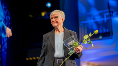 Eine Frau lächelt und steht auf einer Bühne und hält einen Preis aus Glas und eine Rose in der Hand